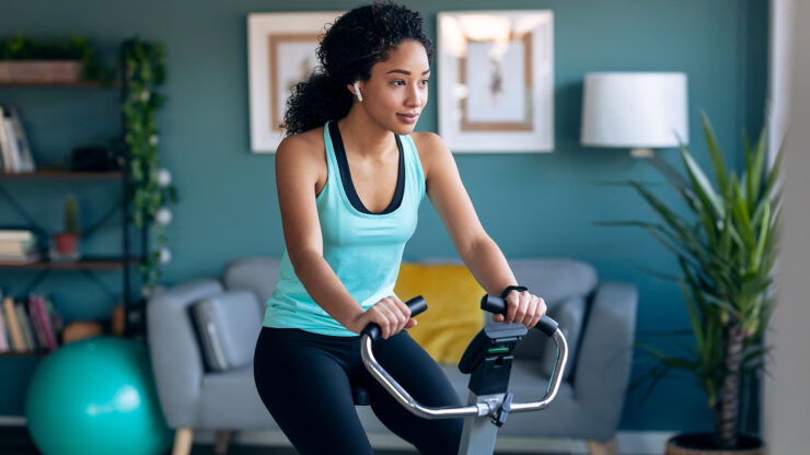 Sporty african young woman exercising on smart stationary bike and listening to music at home.